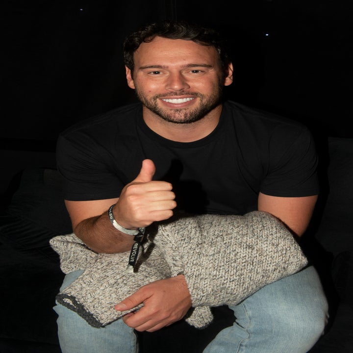 Man sitting, smiling, giving a thumbs-up, wearing a black t-shirt and jeans, holding a knitted blanket