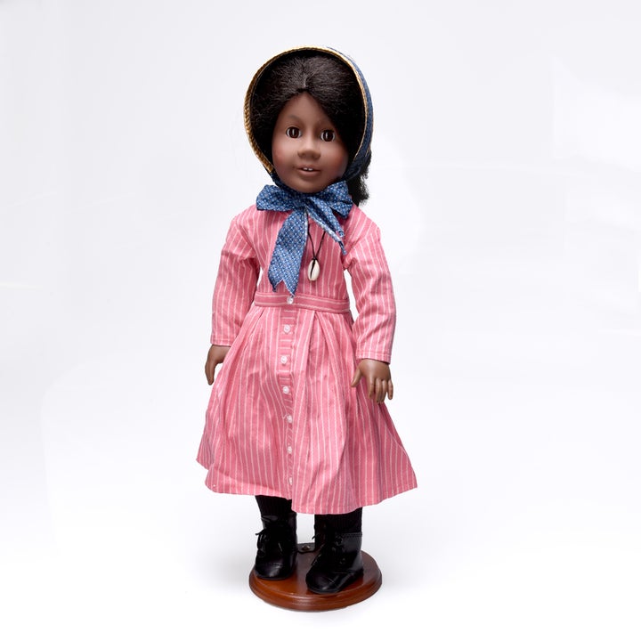 A doll wearing a striped dress, bonnet, and boots stands on a wooden base, styled in a historical fashion