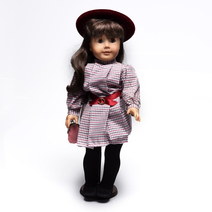 Doll with long brown hair, wearing a checkered dress with red belt and matching hat, black tights, and holding a small bag