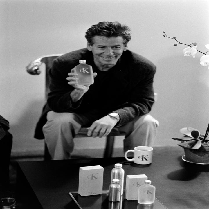 Person sits smiling, holding a CK cologne bottle; other CK products are on the table. 