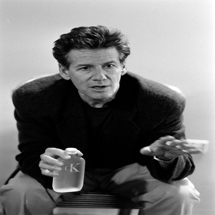 A man sits with a relaxed posture, wearing a casual jacket and holding a bottle with 
